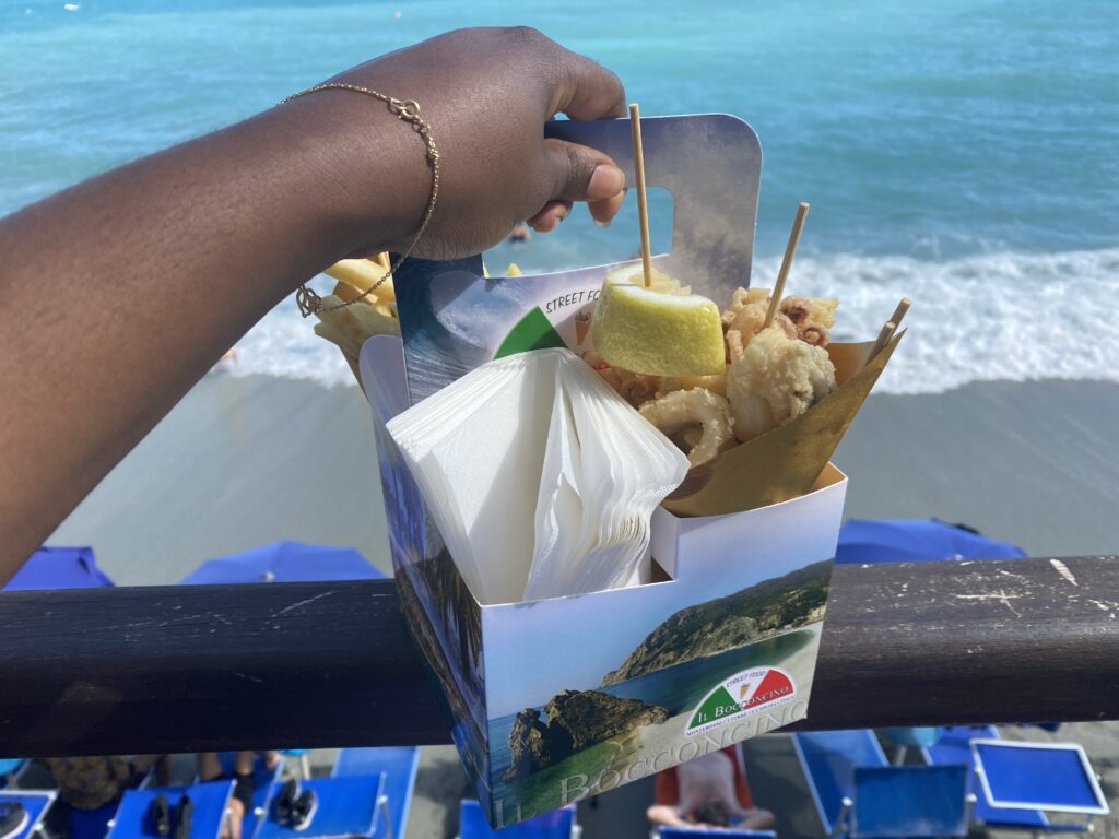 black woman's handing holding a cardbox full of Italian street food. THere is fried fish, calamari, and other fried seafood. 