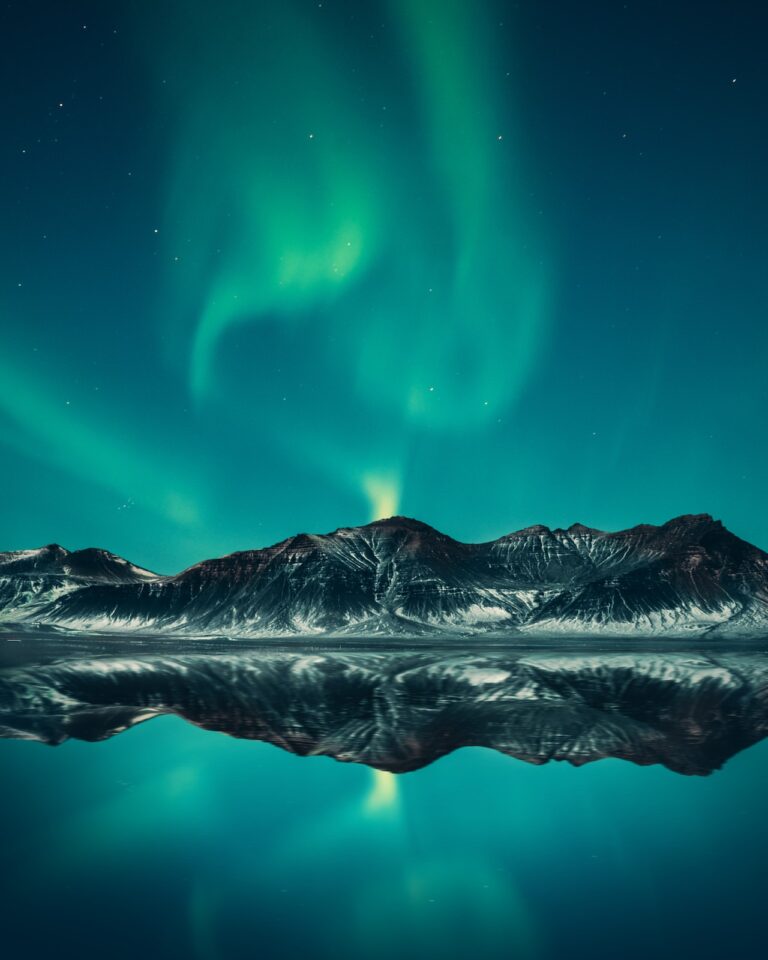 Neon Green aurora lights are above beautiful snowy mountains during the night, in front of mountain is water with reflection of the mountains. I