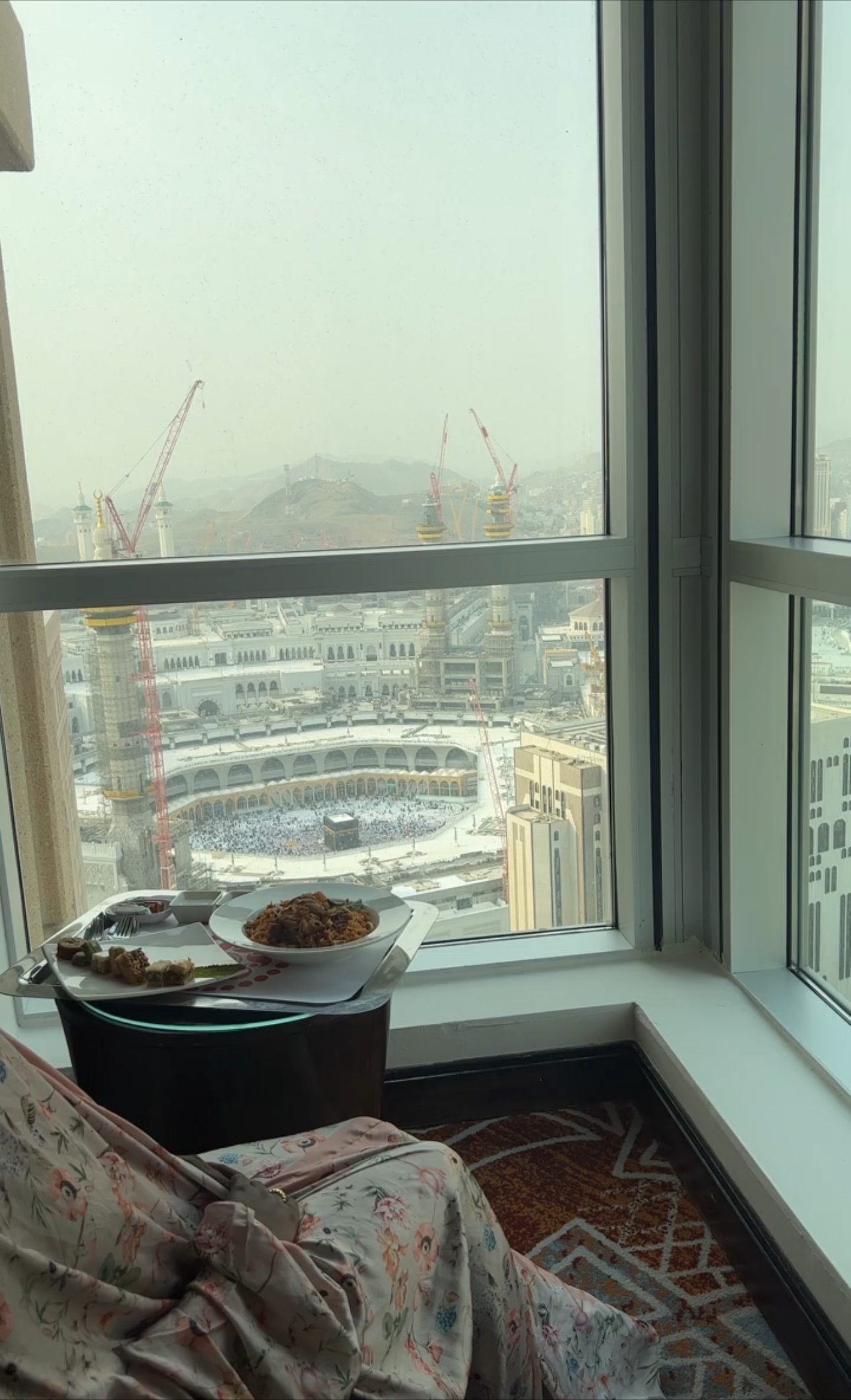Black woman in floral prayer dress eating Arab chicken and rice in her hotel room with a view of the Kaba