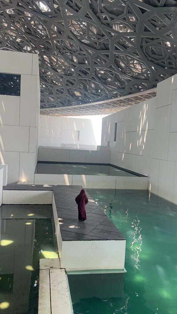 photo of the pool and walkway at Louvre Abu Dhabi. Black woman wearing maroon abaya and hijab walking on runway