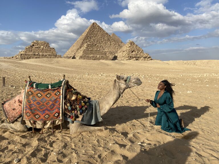 black woman in green dress making a kissy face at a decorated camel in front of the giza pyramids