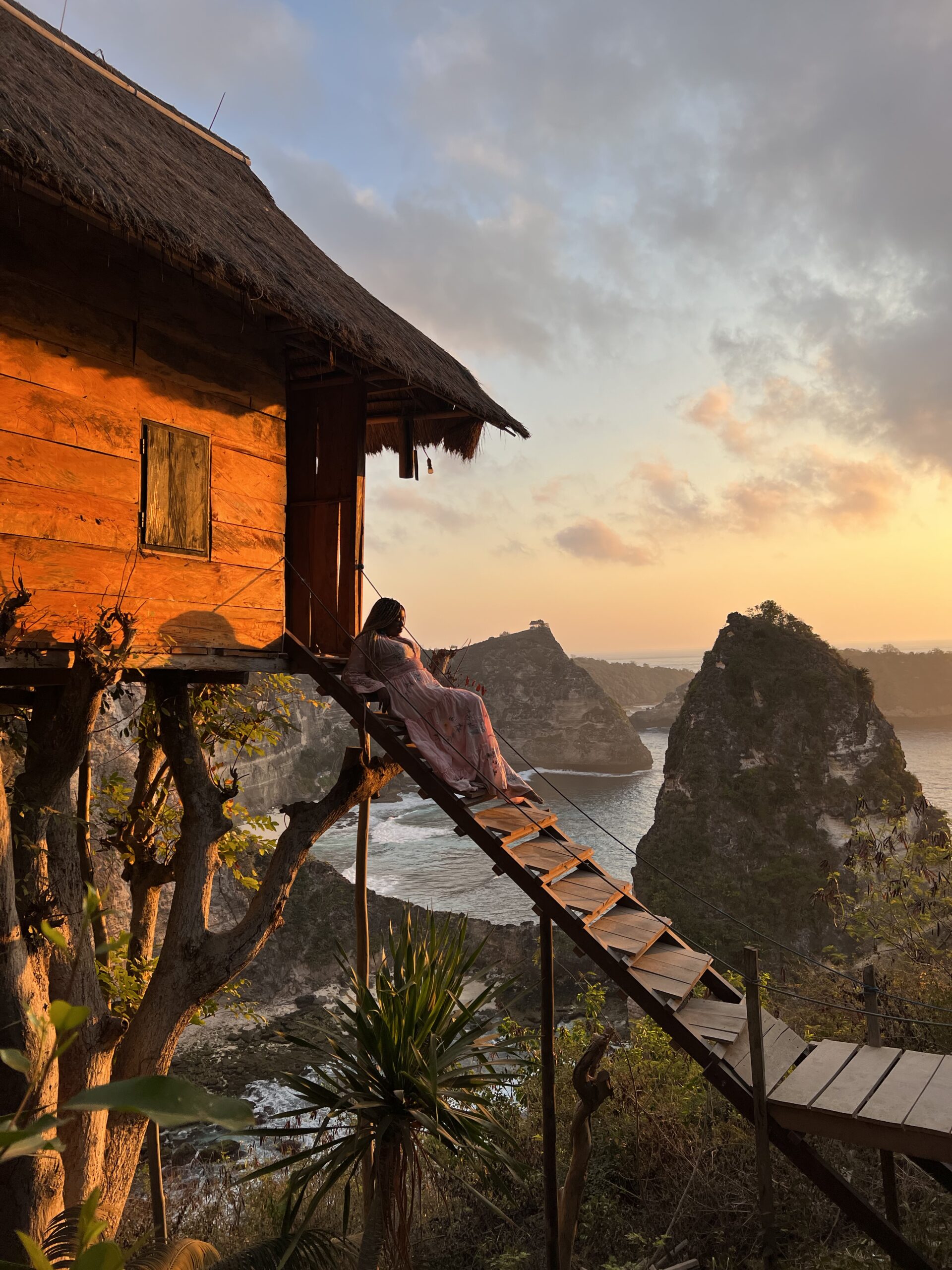 black woman in pink maxi dress sitting on stairs of tree house with a view of the sea. Photo was taken at sunrise and the sky is a bit blue and orange.