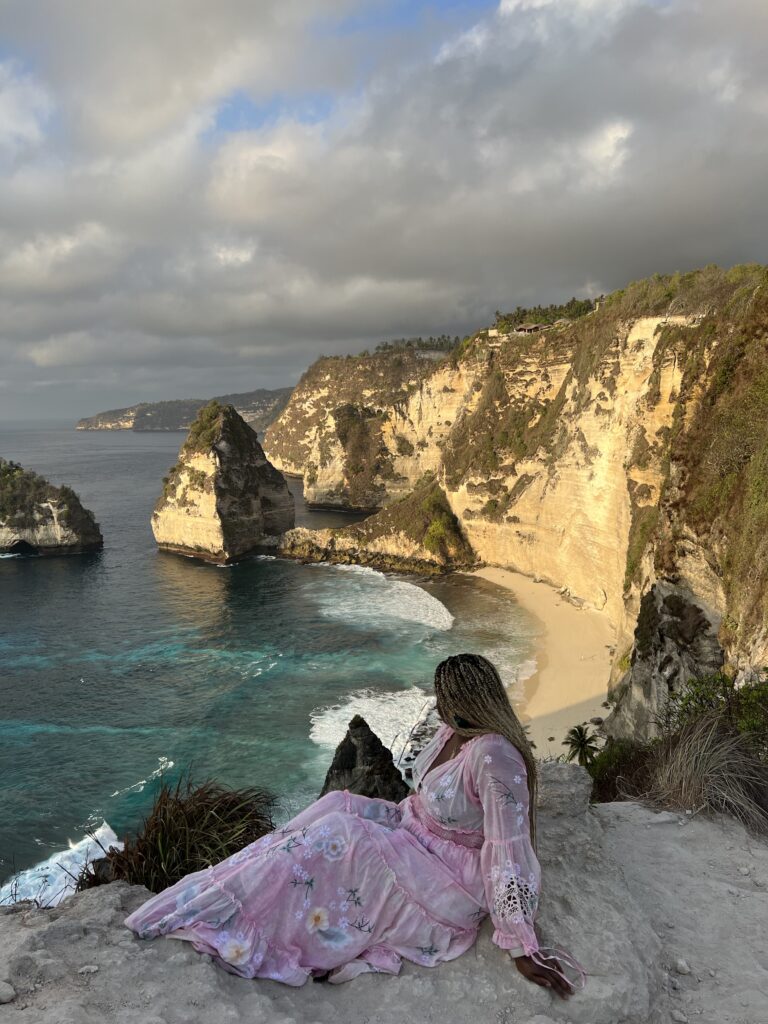 black woman in pink maxi dress sitting in front of rock formations and beach at Diamond Beach 