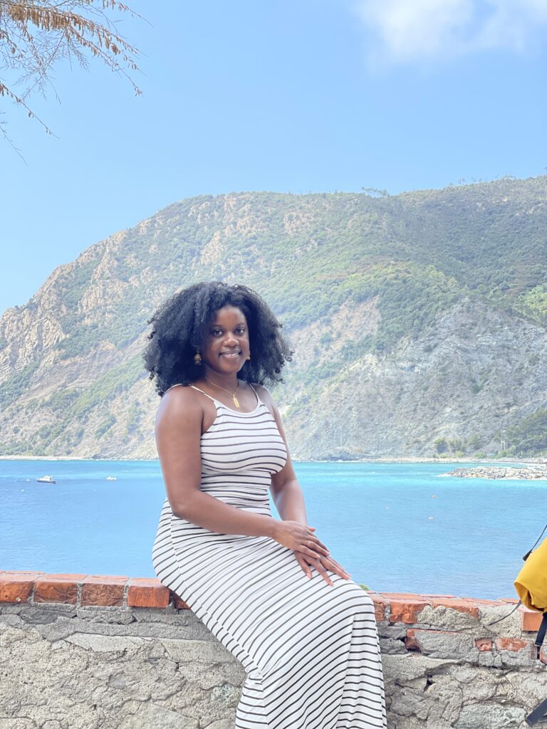 Black woman with afro sitting on stone wall with beach and mountain in the background