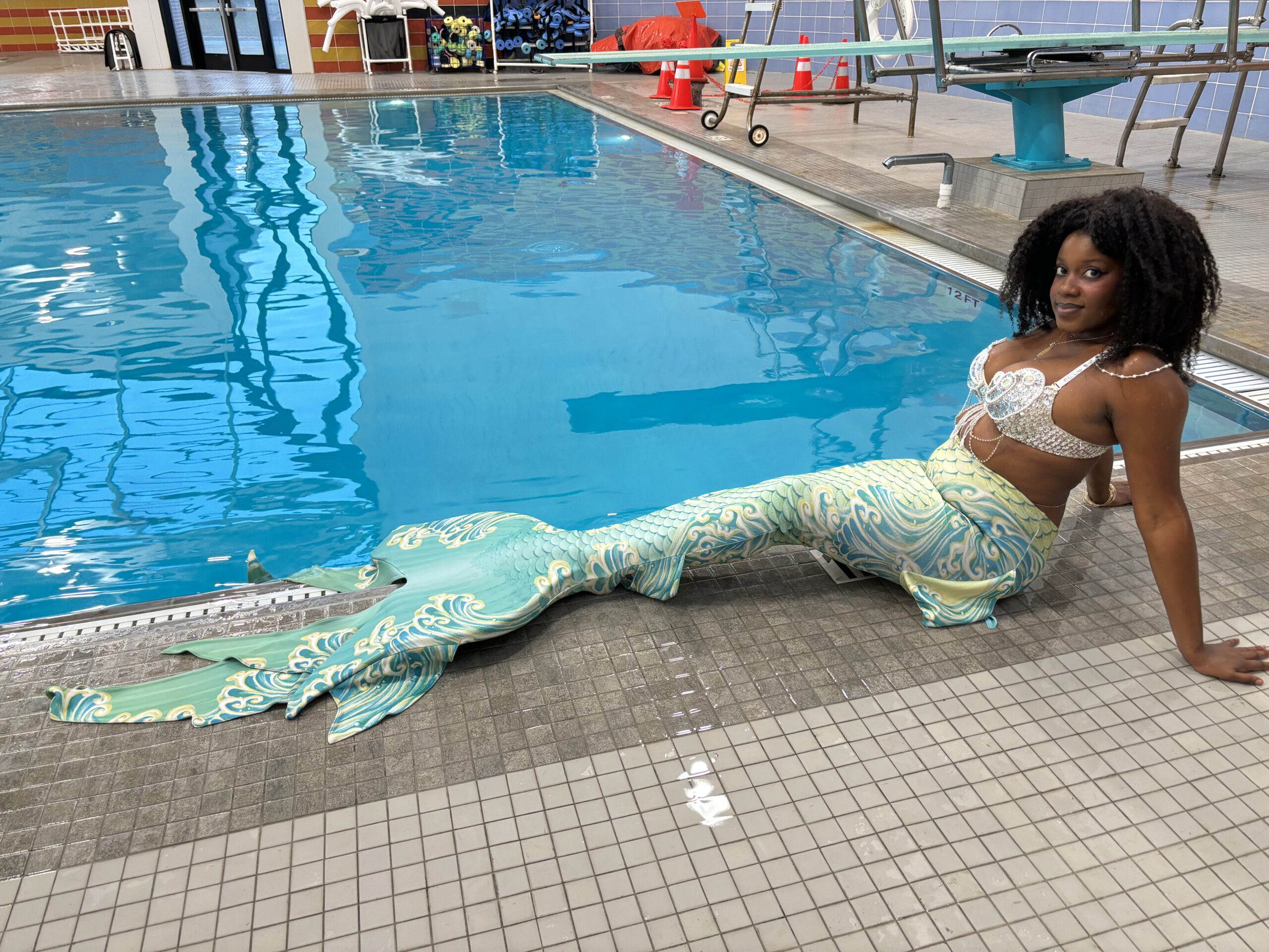 Black woman with afro in green, yellow, and white mermaid tail and silver sequined bra sitting in front of a pool