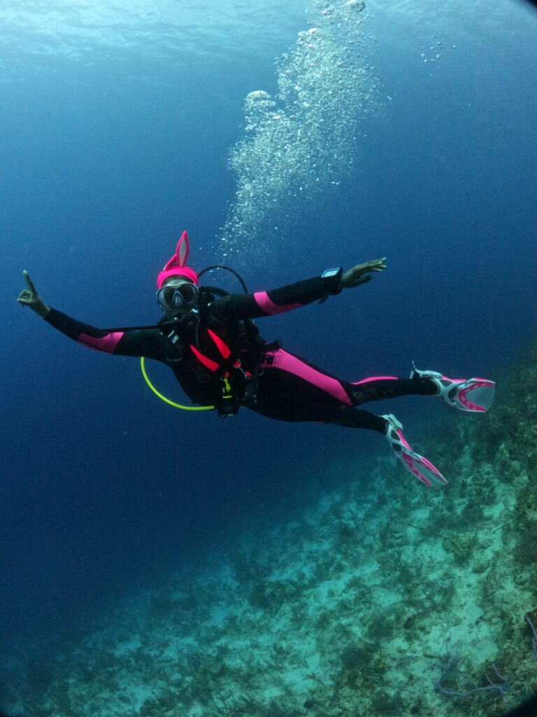 Black woman with bunny ears and pink and black wetsuit giving the peace sign underwater while scuba diving