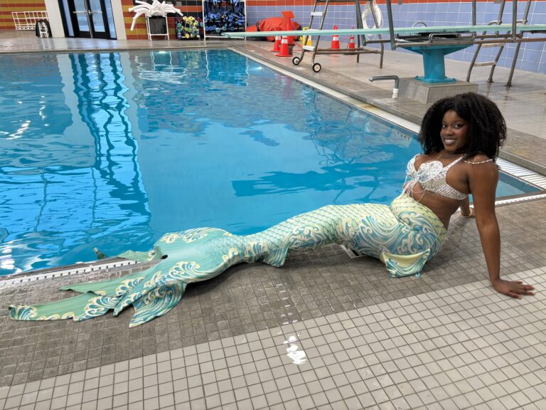 Black woman with kinky-curly hair in green and white mermid tail and white and silver top. She is sitting in front of a pool and smiling.