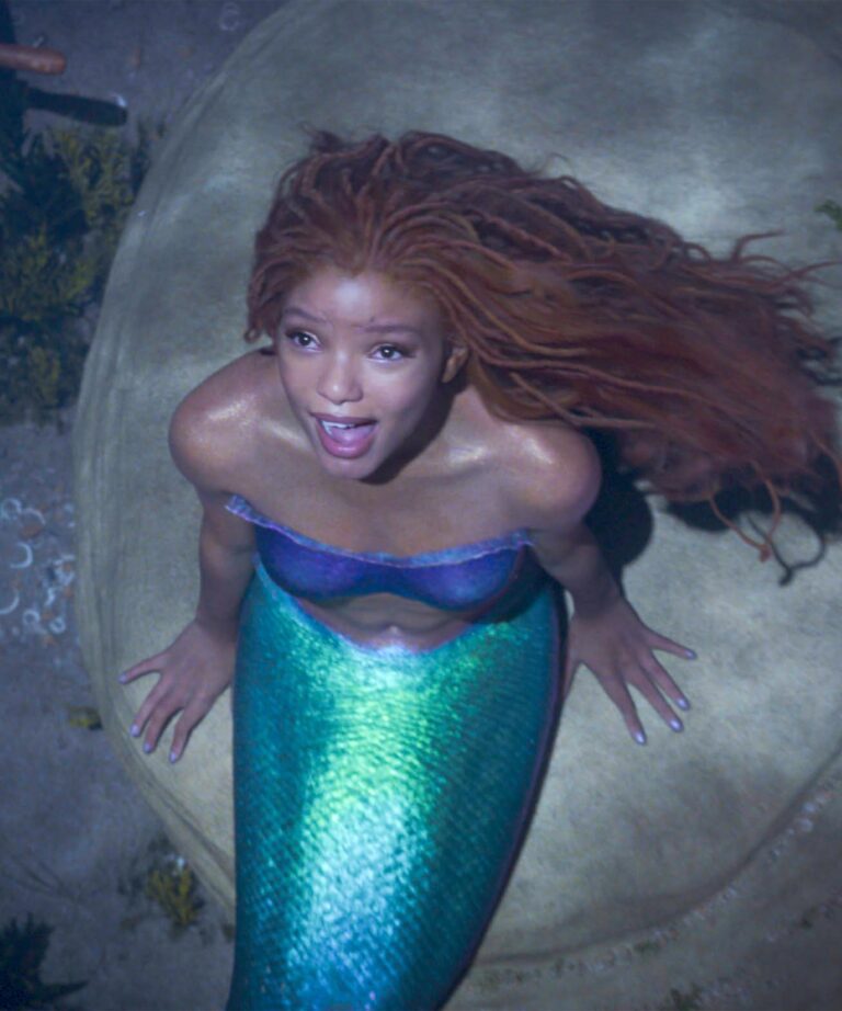 shows Halle as the Little Mermaid. Black mermaid with redish locs sitting on a rock underwater. Wearing a blue top and a green-blueish shiny tail
