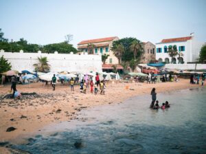 A photo of people and Senegalese style boats (colorful with religious phrases written on them) at the beachfront
