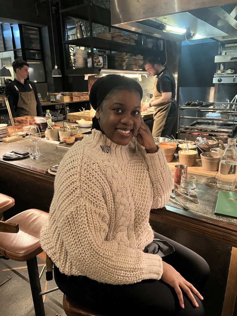 Black woman with black headwrap and beige sweater is smiling at the camera. The woman is sitting at the bar of a busy restaurant 