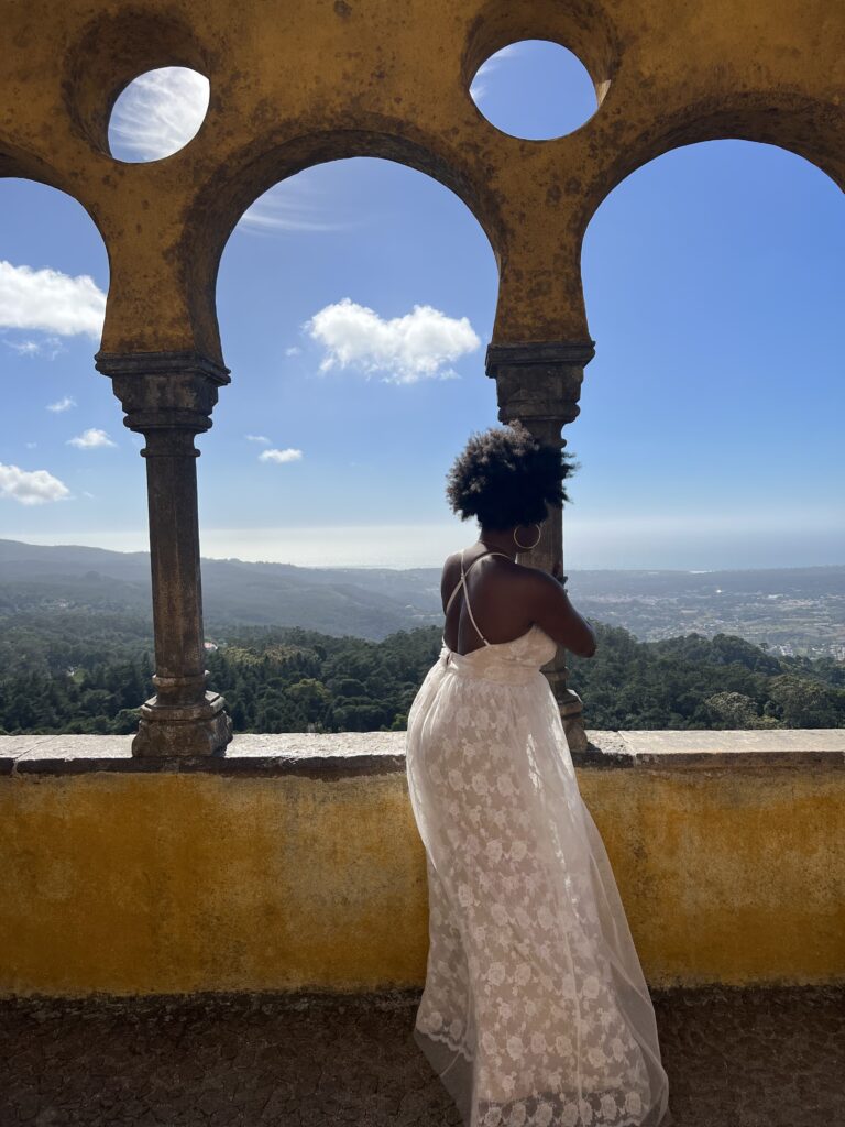 Black woman with afro and white dress in front of yellow walls of Pena Palace with mountains in the background