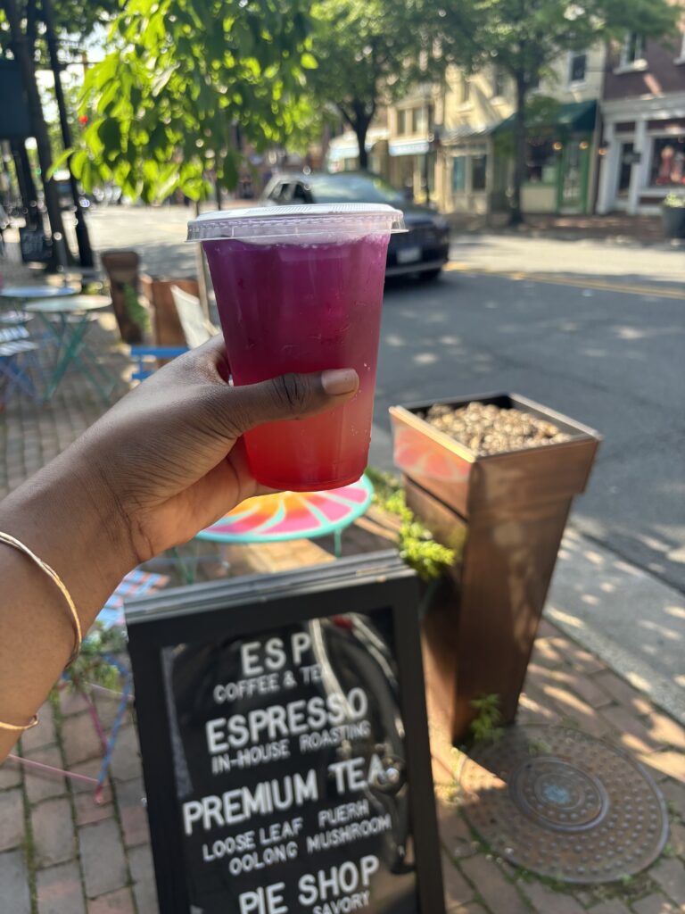 Black woman's hand holding a purple and pink iced drink outside in front of a sign that says ESP coffee and tea