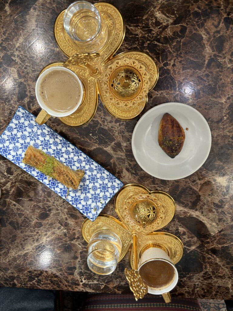 Fancy gold tea sets. One set has milky pistachio turkish coffee. The other set had dark brown coffee. Turkish pistachio pastries, water, and turkish candie are also on the table.