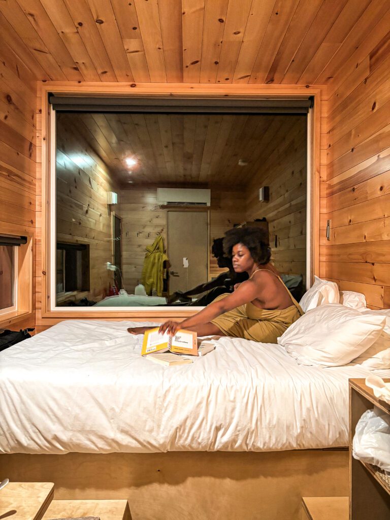 dark skinned black woman with black afro in olive green dress sitting on a bed with white sheets, reaching towards a book. In front of a big window that takes up half the wall. Located in a tiny Getaway House cabin in the woods
