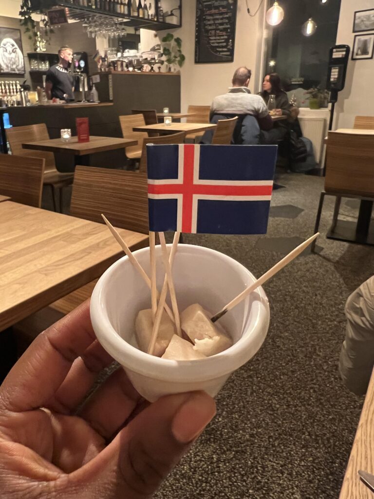 black woman's hand holding cut pieces of hakarl or fermented shark with mini flags of iceland toothpicks