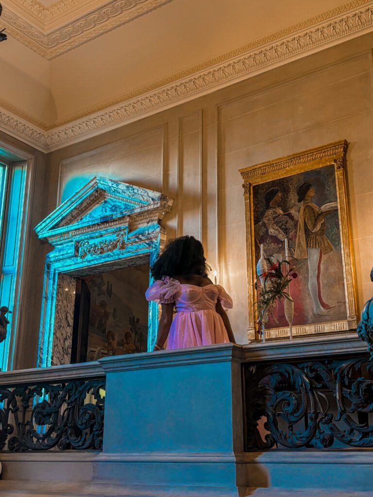 Dark skinned woman with afro and pink dress standing in front of medieval era painting on top of a staircase 