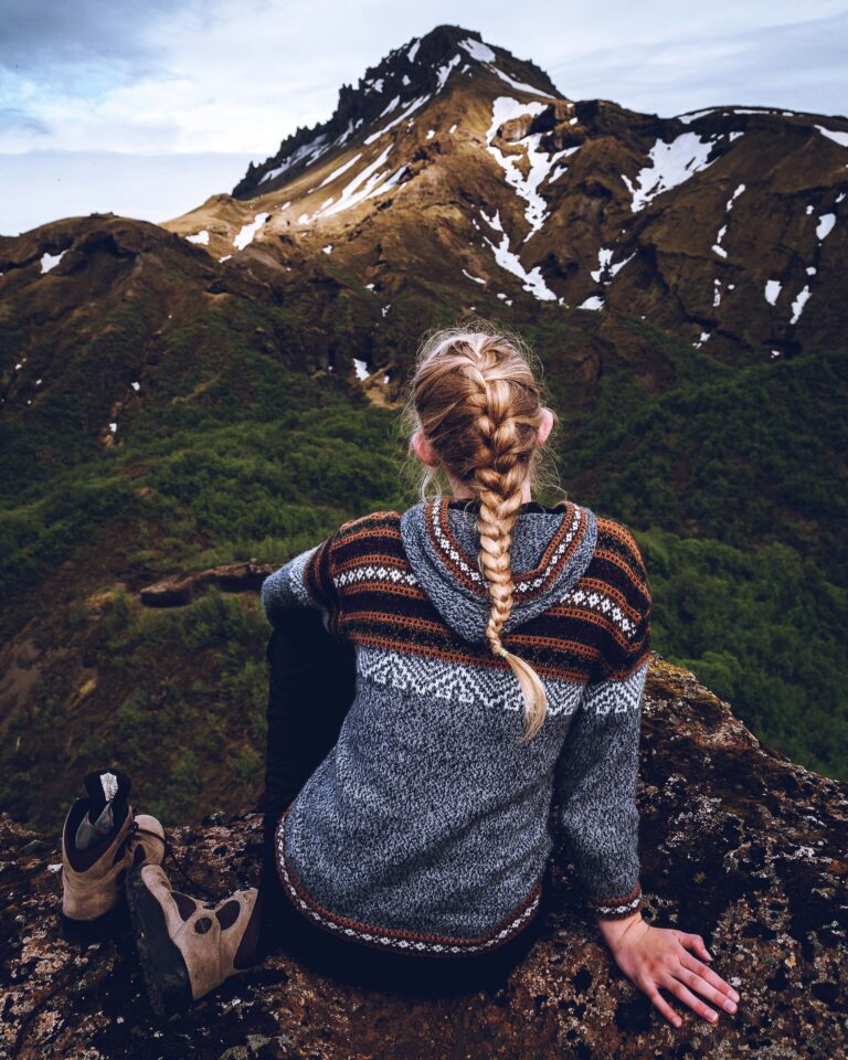 Blonde woman with long braid wearing a traditional Icelandic wool sweater called Lopapeysa. She is sitting on a rock facing a mountain with slight snow and greenery.