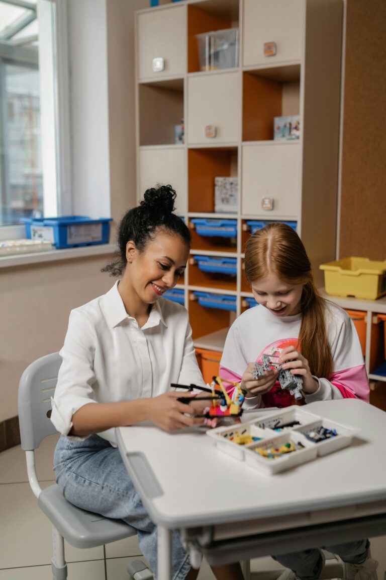 Light brown skinned woman with slight curly hair and pale skinned child with red hair playing with legos at a tale in a classroom.