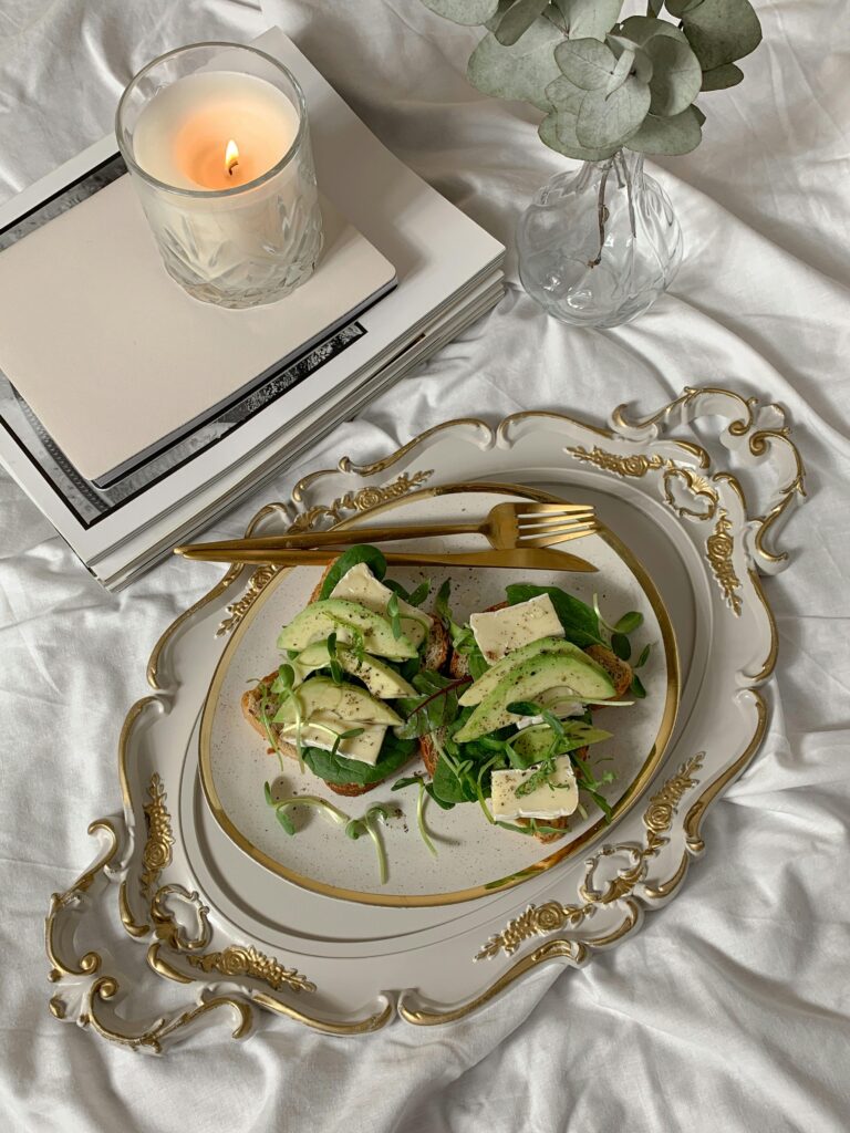 breakfast in bed, avocado toast on fancy gold and white plate. A lit candle is next to it. The sheets are white