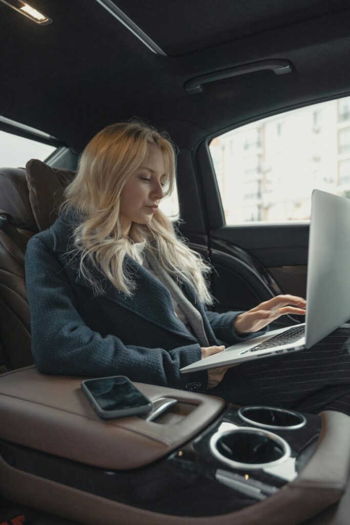 white woman with blonde hair sitting in the back of a fancy SUV, typing on her laptop