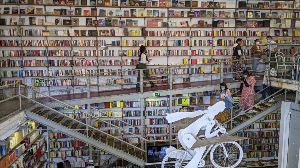 A bookstore that has floor to ceiling books. Shows a Black woman with an afro puff and wearing black combat boots walking in front of books.  There's a wooden sculpture of a person riding a bicycle with their scarf swaying in the air in the center of the staircase. 