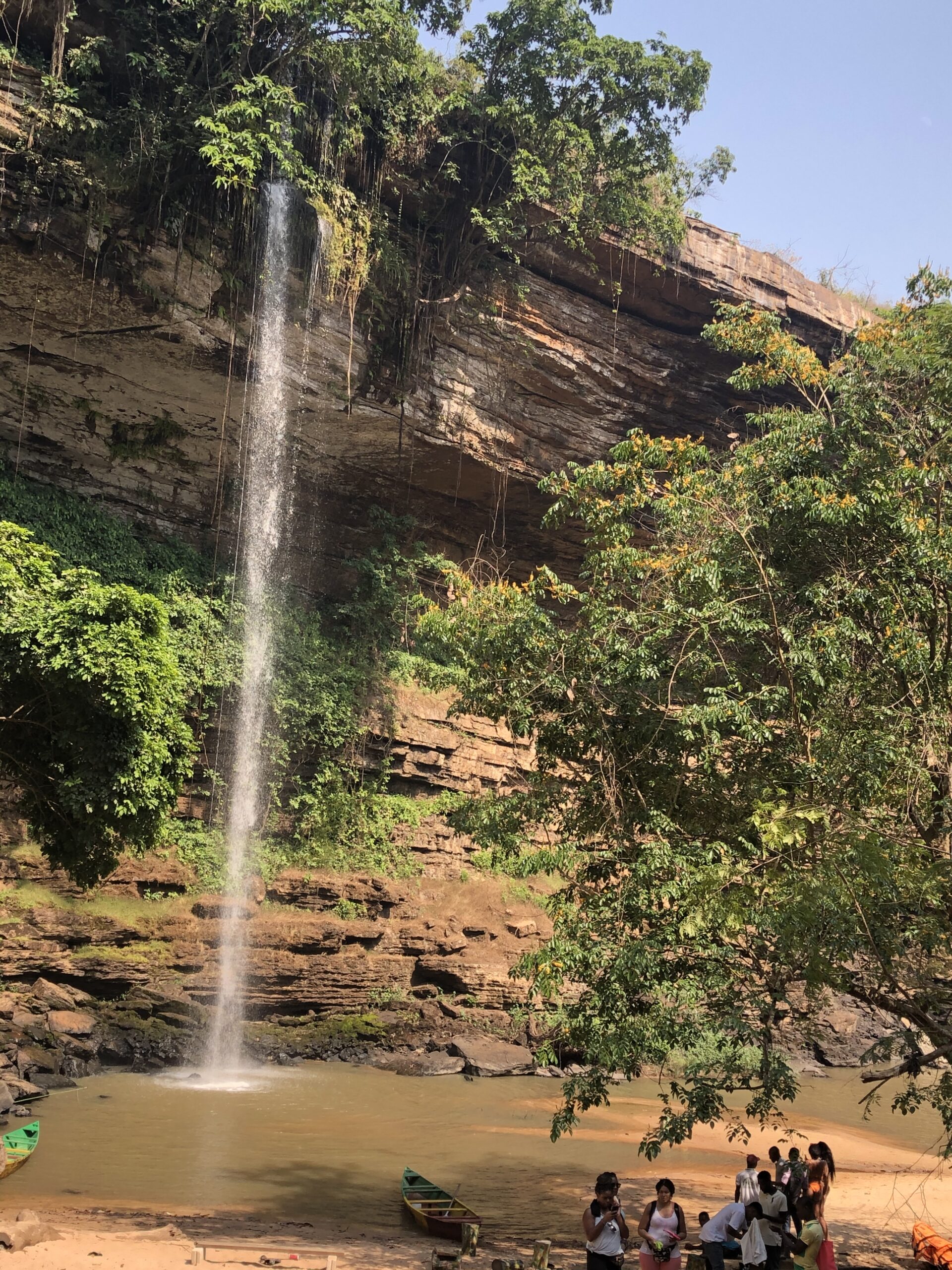 Tall waterfalls at Boti Falls that causes a small ravine. Small green canoe at the bottom with lots of Black visitors at the bank of the ravine.
