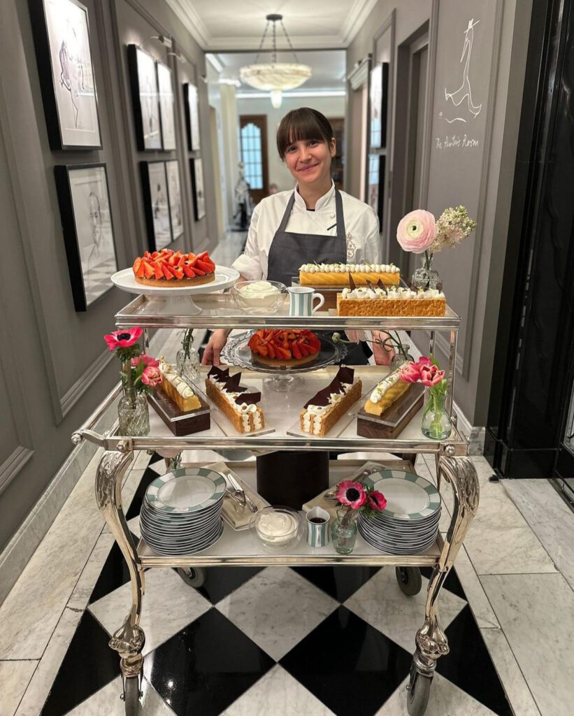 a white female chef or waitress with brown hair posing in front of trays of afternoon tea pastries and floweres