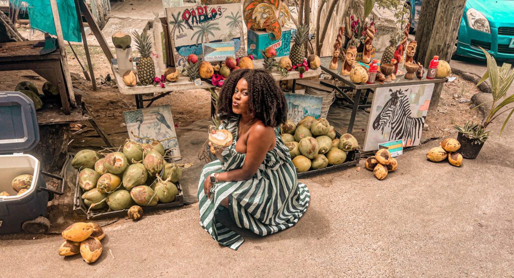 Dark skin woman with afro wearing an African green and white stripped dress, she is smiling and holding a coconut next to her mouth. In the background is a fruit vendor with lots of coconuts and other tropical fruits. 