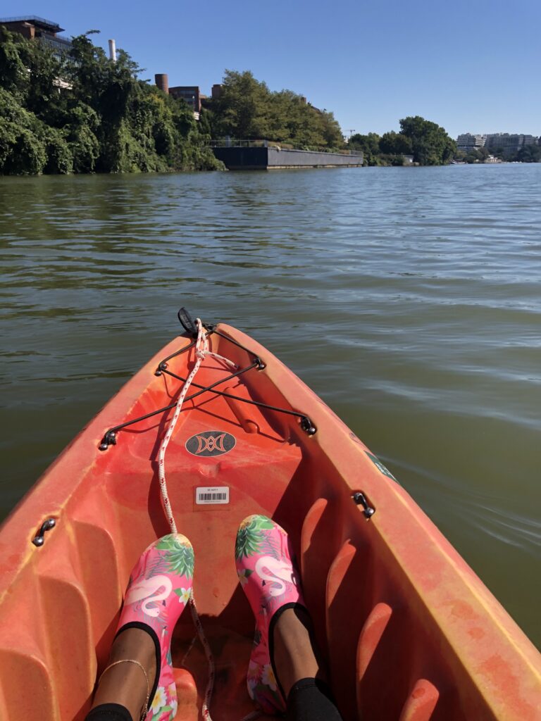 dark skinned legs in flamingo water shoes in a orange kayak. The bathground is green-brown fresh water with building far in the distance