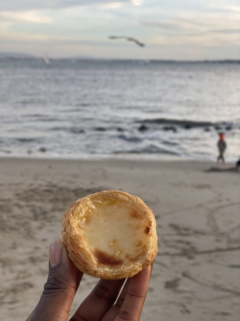 dark skinned hand holding a pastel de belem with a beach in the background. There's a kite flying in the background