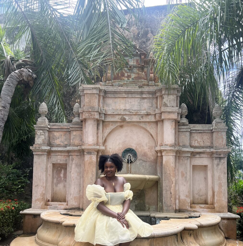 a dark skin Black woman in a yellow princess dress sitting in front of Fuente Paseo La Princesa (a water fountain) 