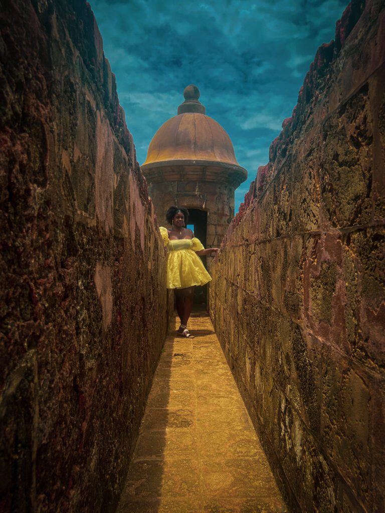 Dark skinned woman in yellow princes style dress and afro hair standing in front of a turret at Castillo San Felipe del Morro