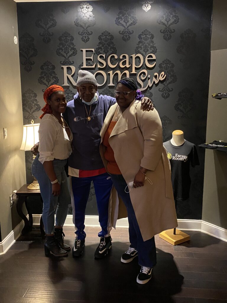 2 dark skinned Black women and an older Black male with Escape Room Live written on the wall 