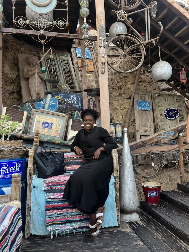 Black woman with afro puff and black dress sitting in front of eclectic furniture and bicyle decor