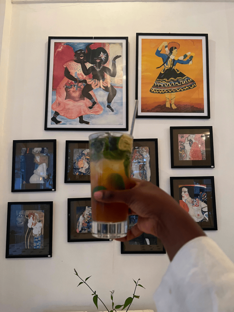 Black woman's hand holding a mocktail that's brown and yellow with mint leaves. The background includes various pieces of art depicting women of all colors dancing. 