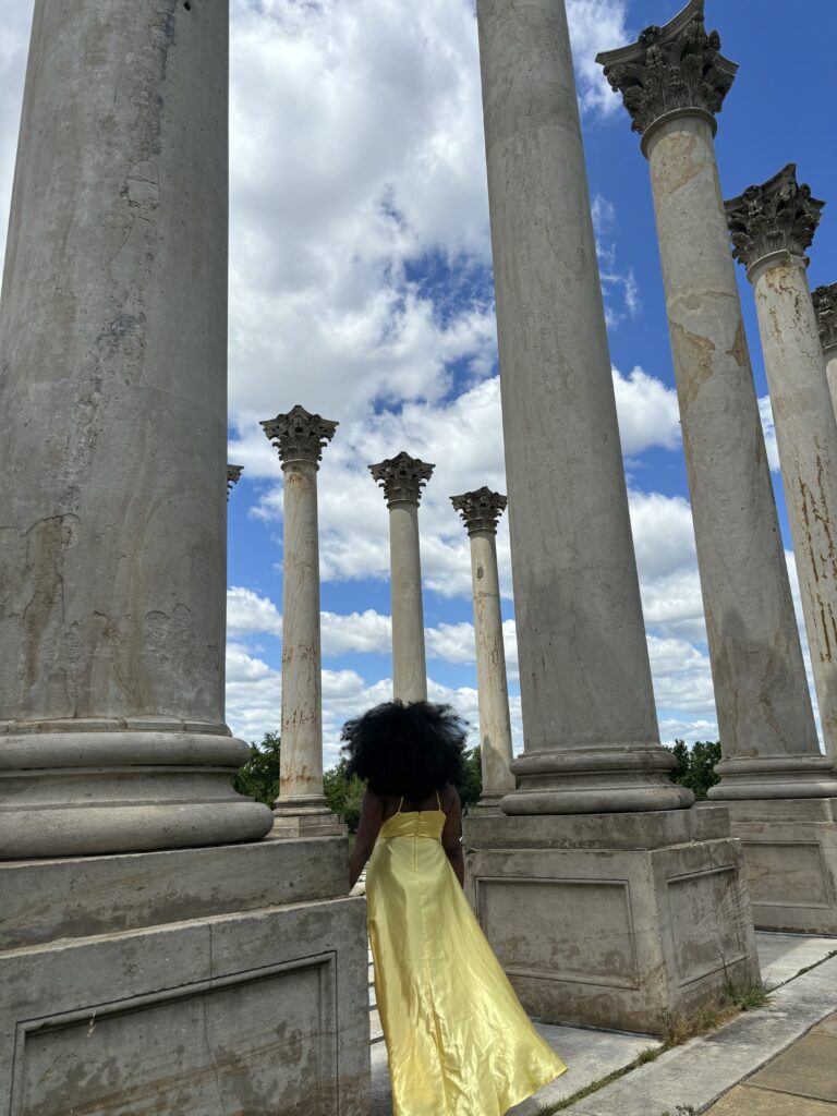 Black woman with long afro in yellow dress standing next to a large column with other columns around her