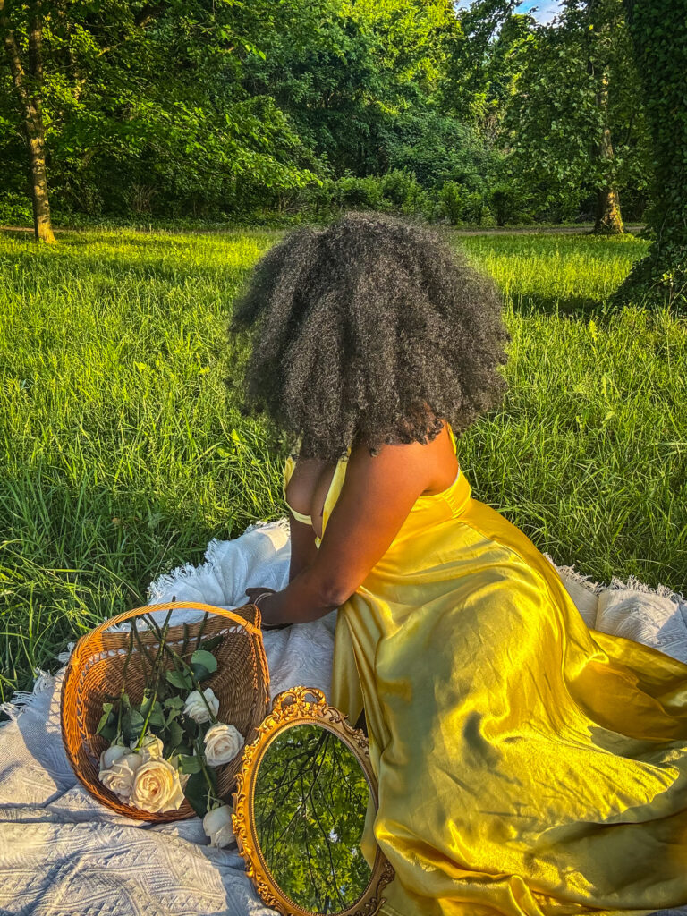 black woman with large afro in yellow dress laying on white sheet next to a tree. On sheet are  flowers ,flower basket, and vintage gold mirror.