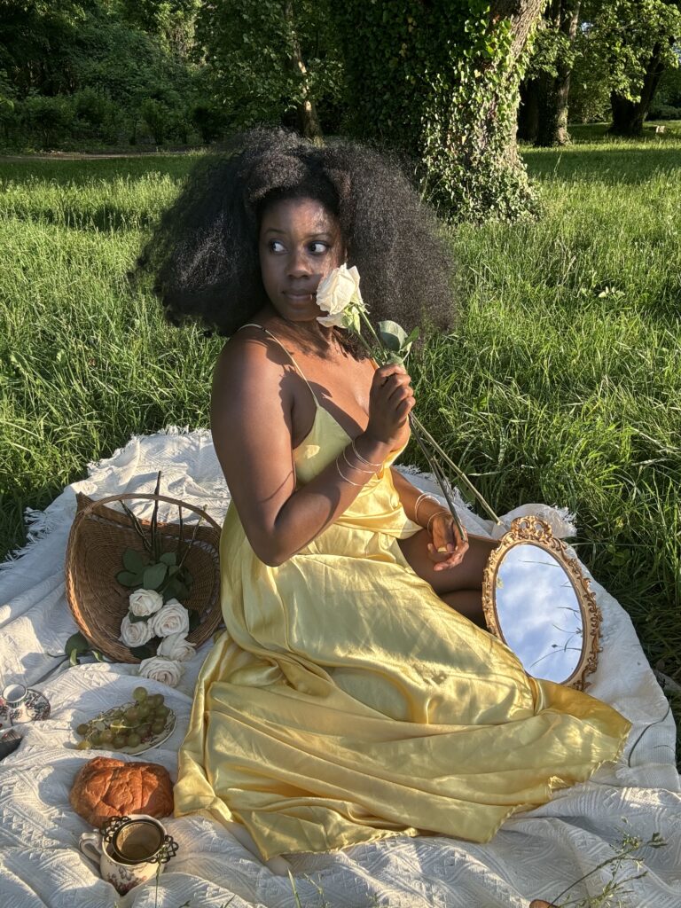 a Black woman in a yellow dress and large afro smelling a flower while having a picnic. 