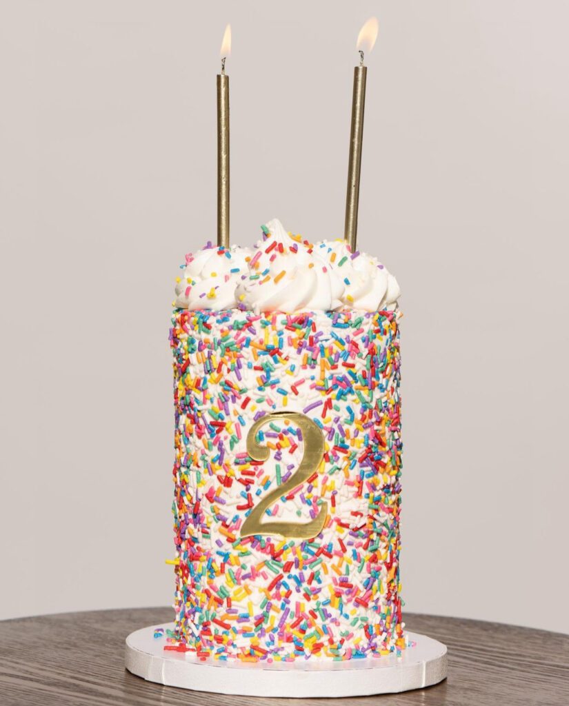 tall cake with color funfetti sprinles and two candles on top. A gold number 2 is on the front of the cake