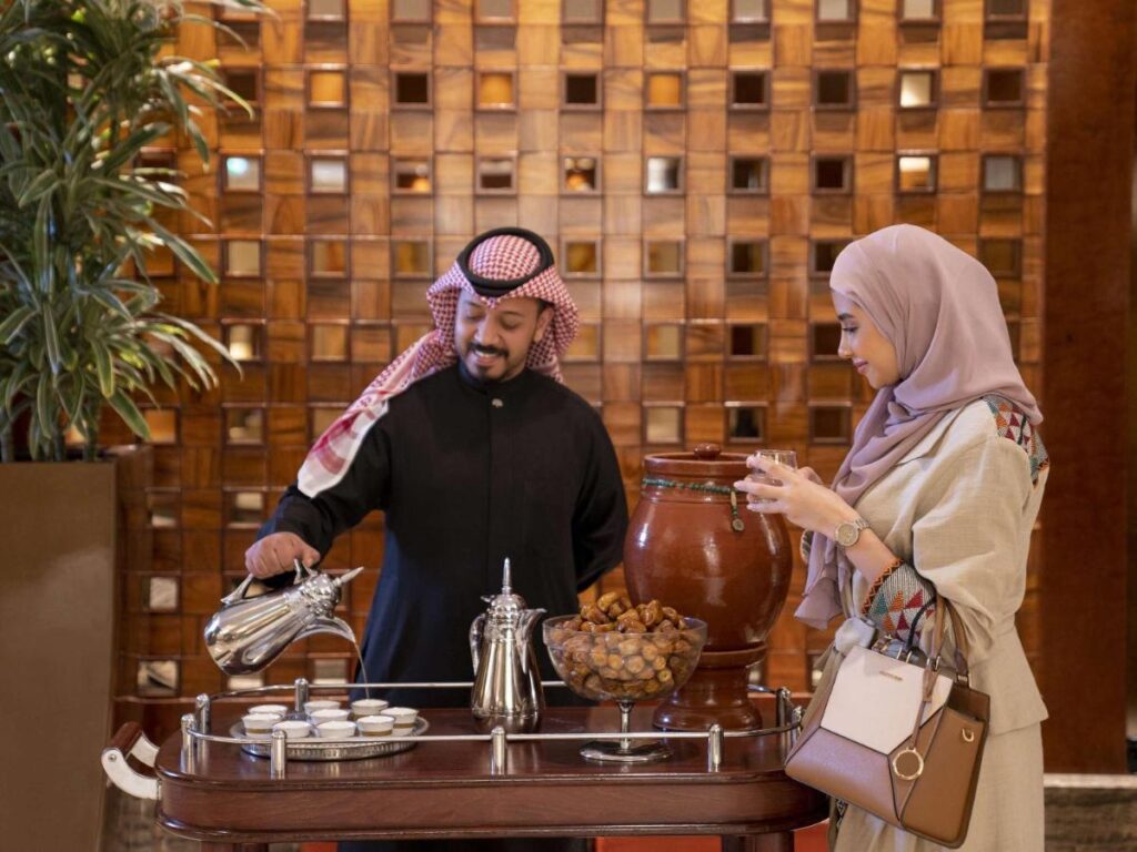A man wearing Saudi headwear pouring coffee for a woman wearing a hijab and abaya 