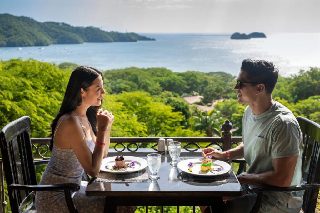 a woman and man are enjoying a meal at a table overlooking the mountain and ocean.
